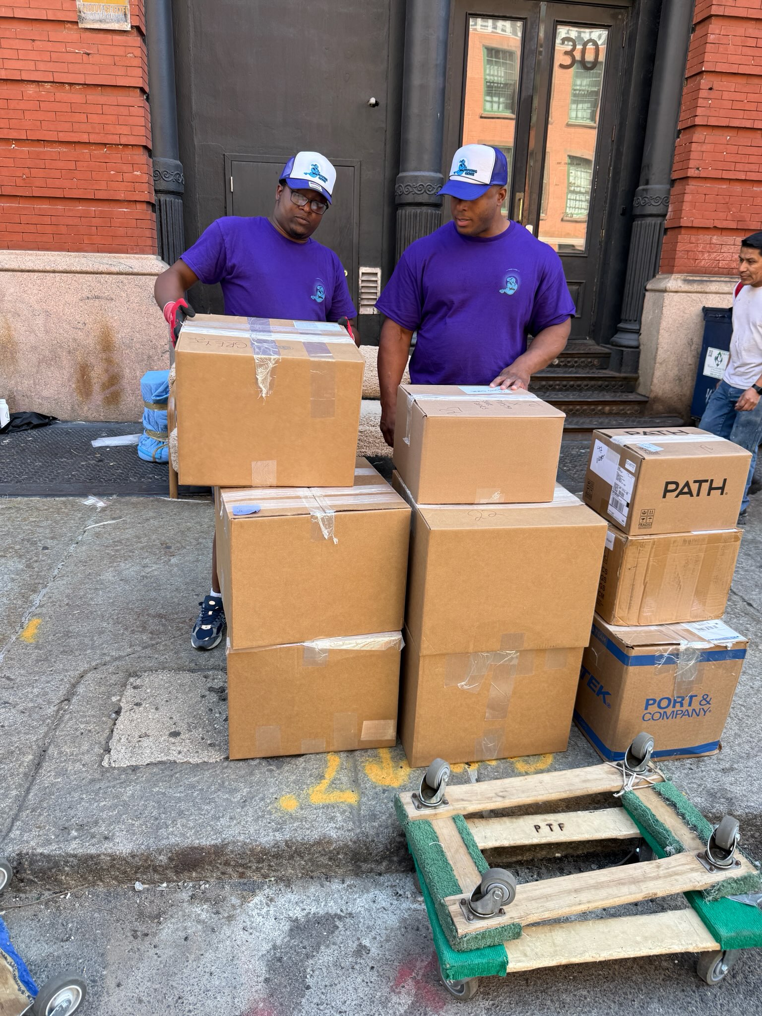 Two Moving Genie movers in branded purple shirts and caps with a stack of packed boxes and a furniture dolly on a Manhattan street