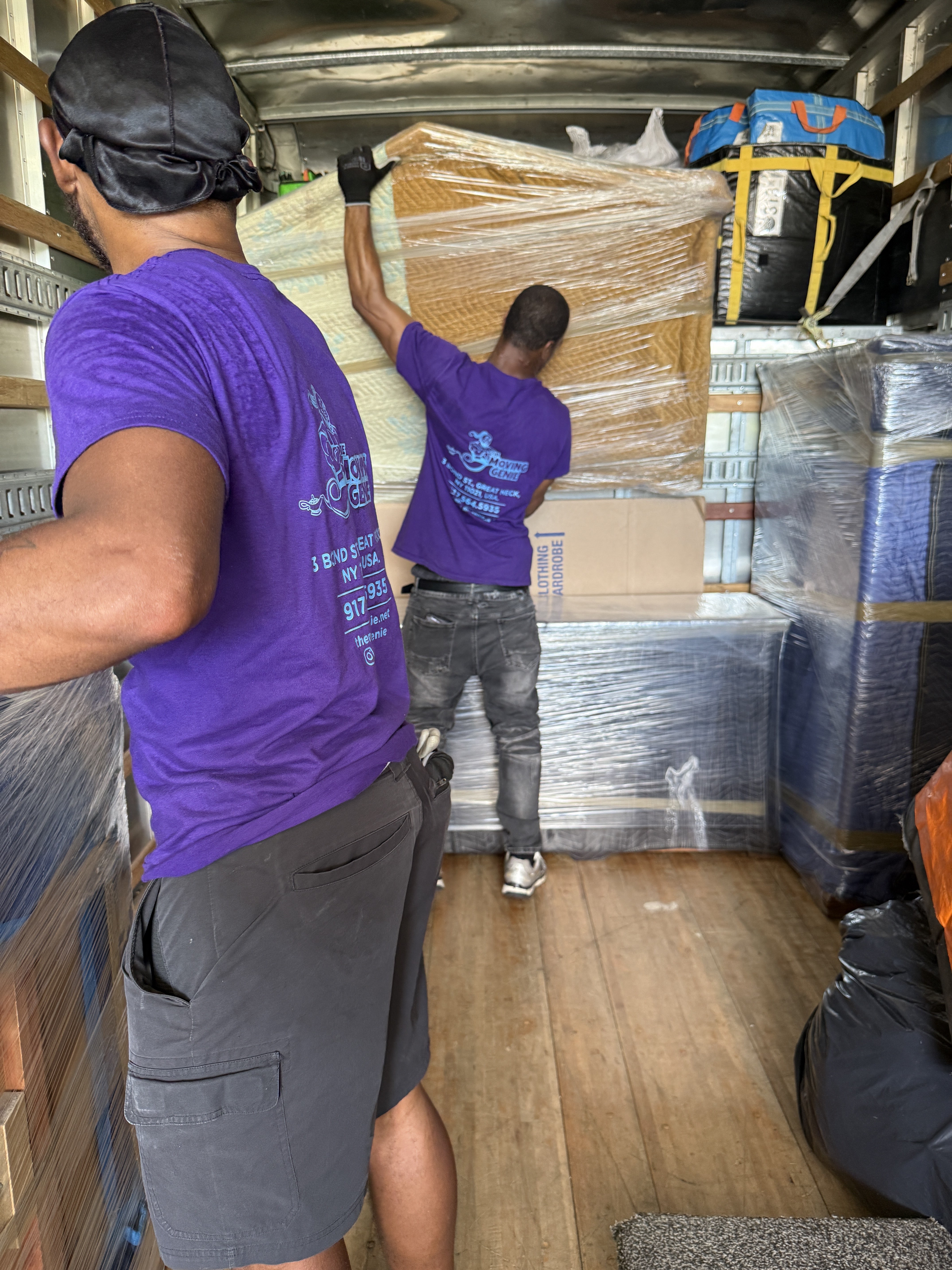 Two Moving Genie movers in purple shirts loading and stacking wrapped furniture inside a moving truck