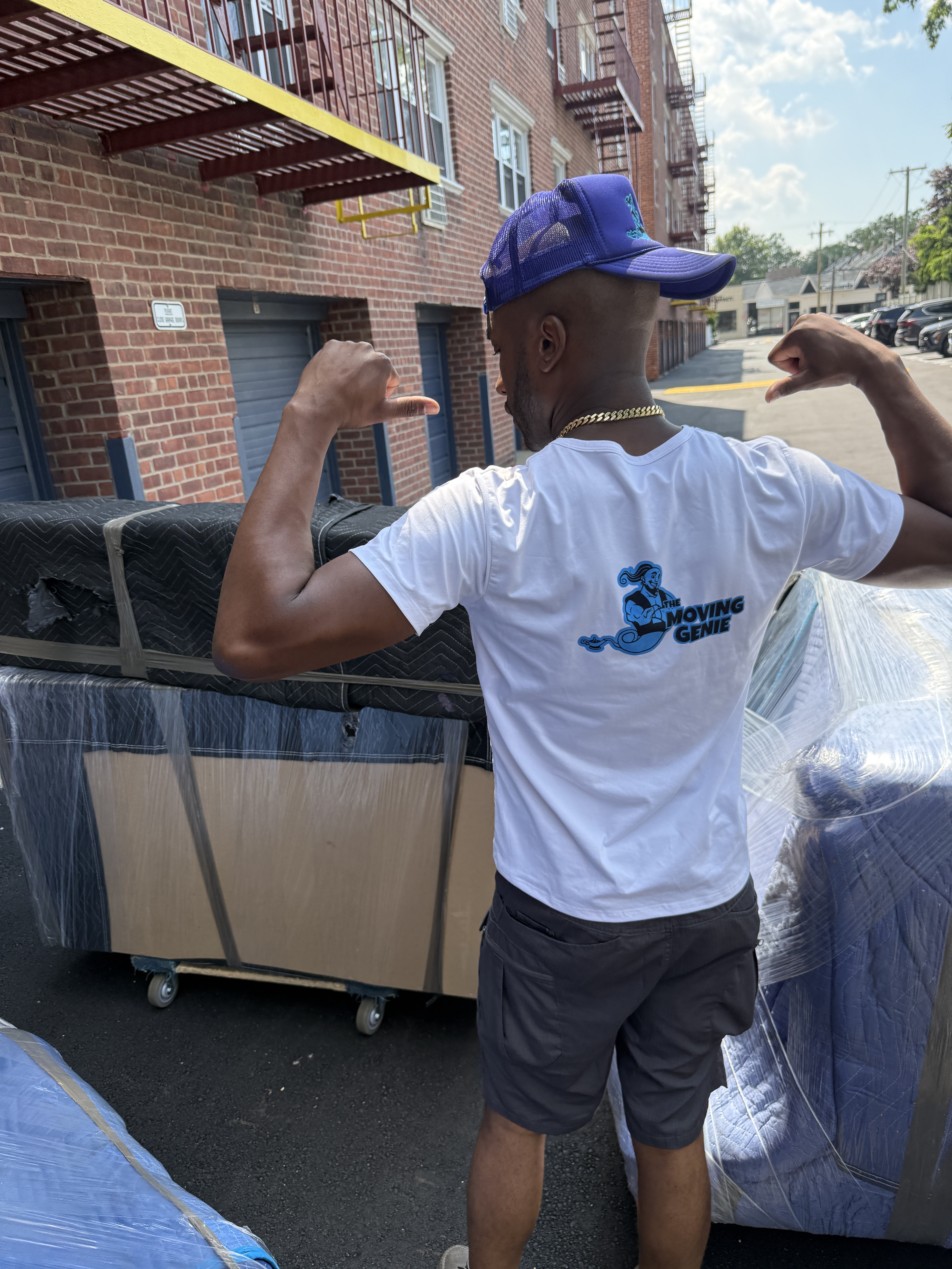 A Moving Genie crew member in a branded shirt beside wrapped furniture on a loading dolly outside a Queens apartment building