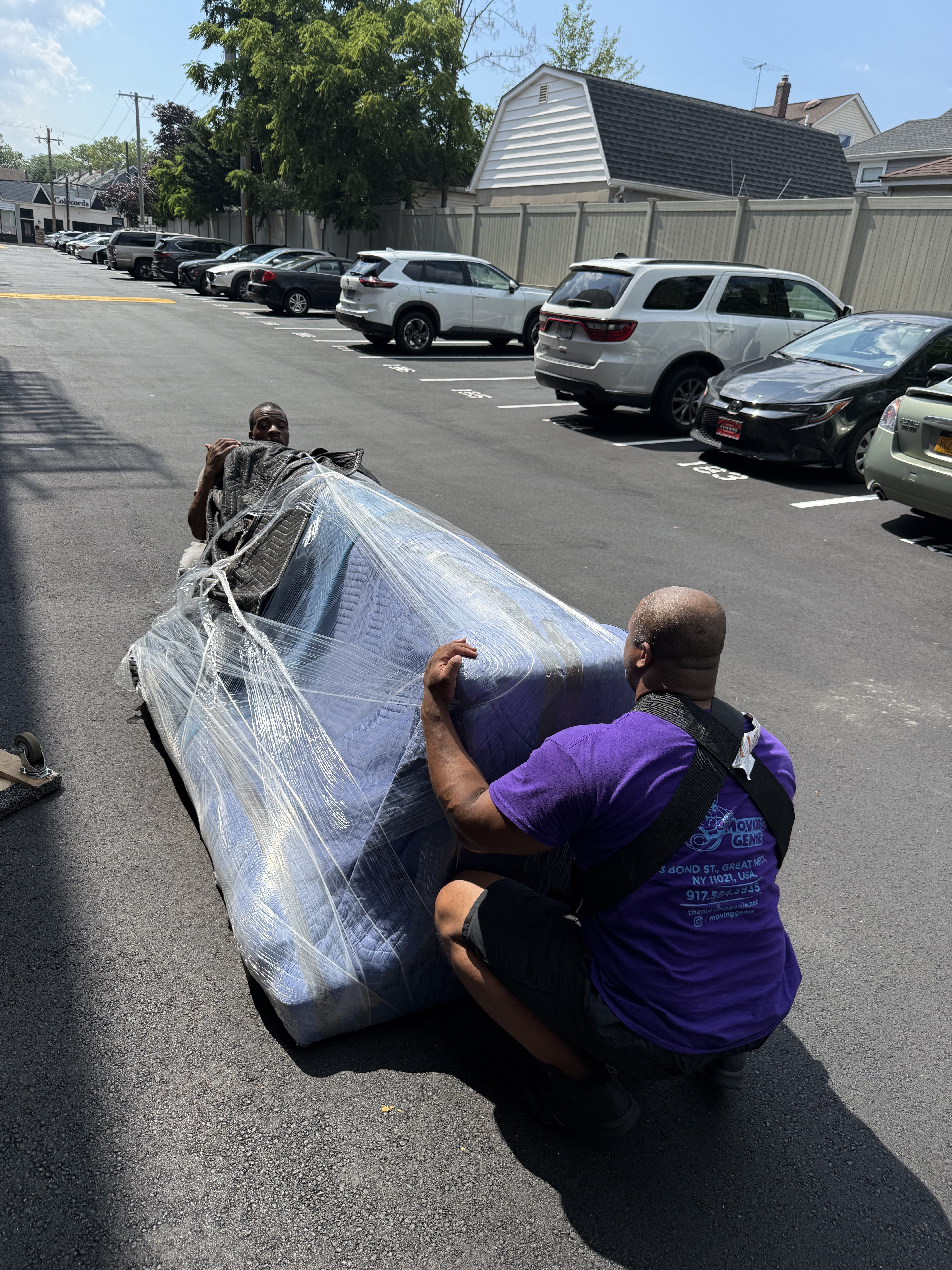 Two Moving Genie movers in purple shirts blanket-wrapping a sofa in a Nassau County parking lot