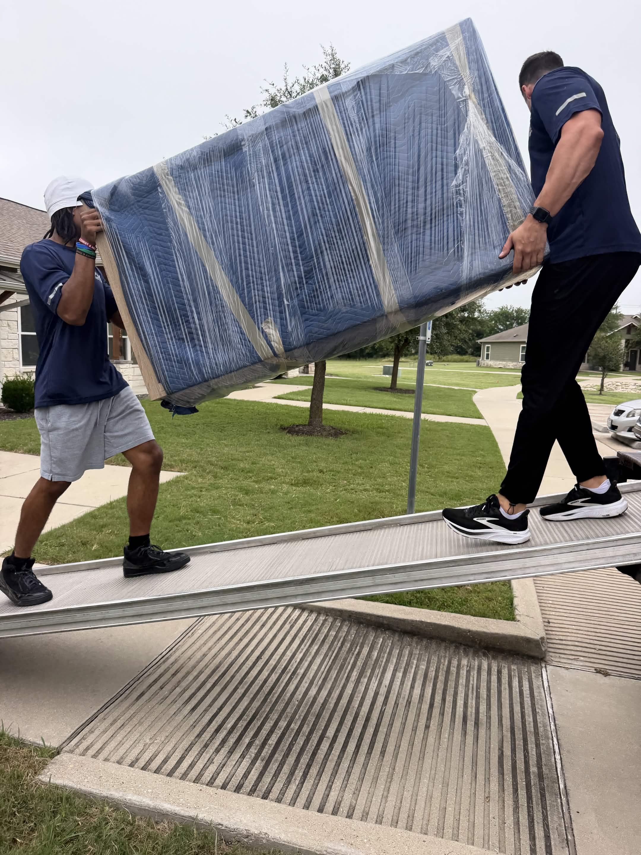 Two Playhouse Moving crew members carrying a shrink-wrapped sofa down a truck ramp in a suburban Austin neighborhood