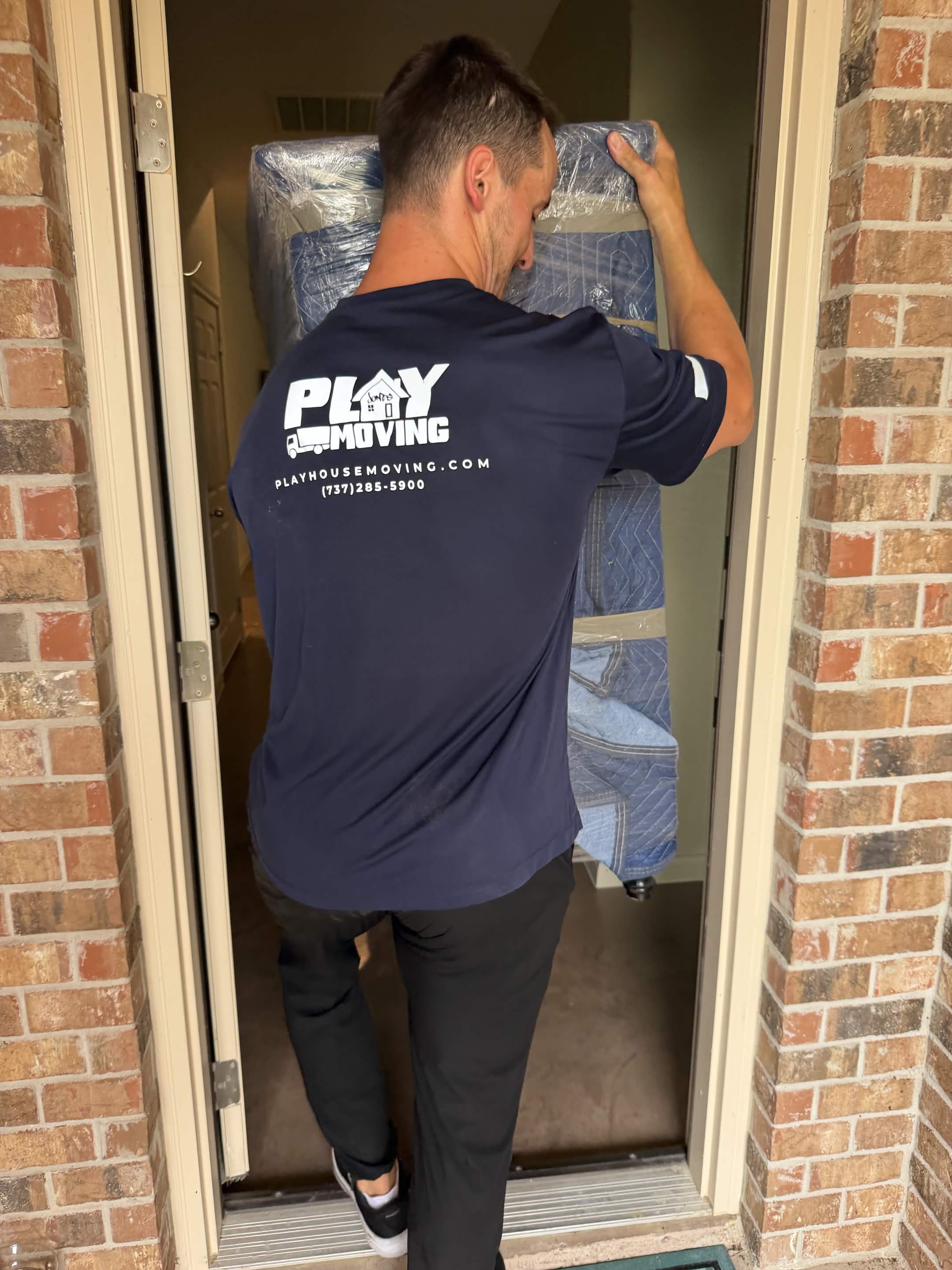 A Playhouse Moving crew member in a branded shirt carrying a shrink-wrapped mattress through a home entryway